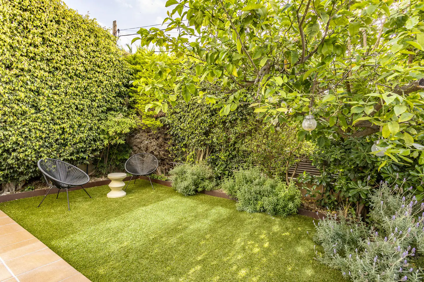 A cozy backyard with artificial grass, two black chairs, a small table, and lush green foliage.