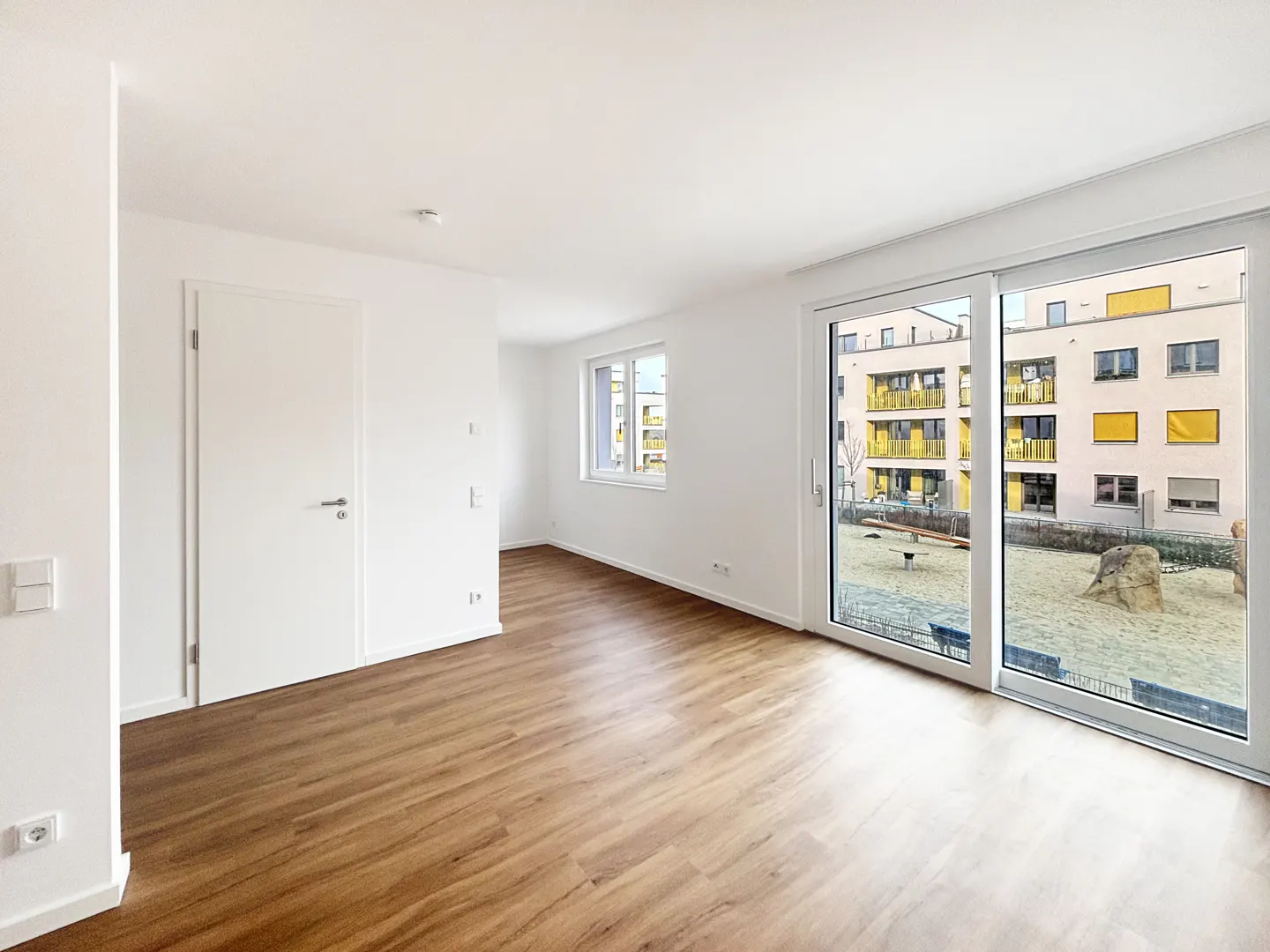 Bright, empty room with wood-look floors, white walls, and large sliding glass doors overlooking a playground.