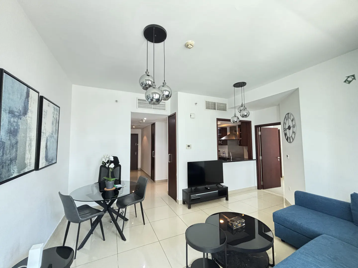 Bright apartment interior with a glass dining table, blue sofa, and modern lighting fixtures. Neutral tile flooring and white walls create a spacious feel.