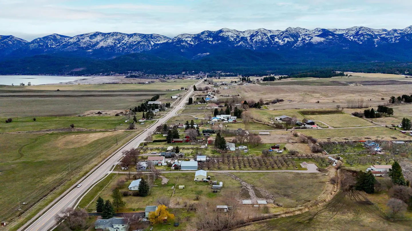 Aerial view of a rural town with a long road, houses, fields, and snow-capped mountains in the background.