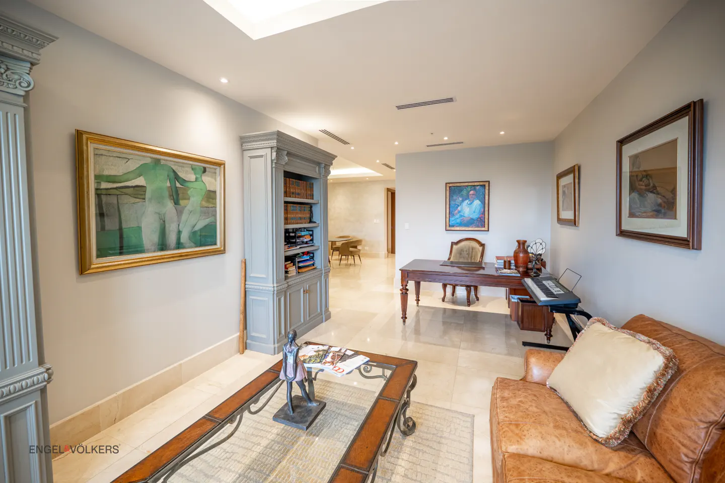 A well-lit living room with a brown leather sofa, glass coffee table, desk, and artwork on the walls.