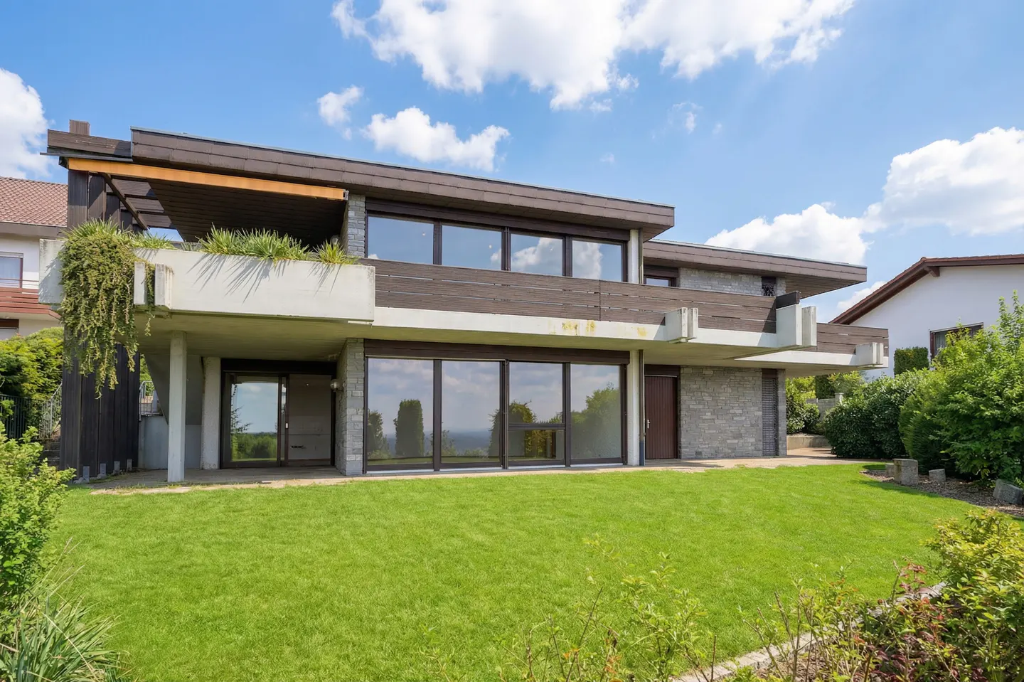 Two-story house with a flat roof, stone facade, and large windows, surrounded by a green lawn and blue sky.