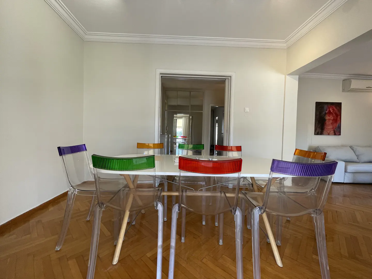 Bright dining room with white walls, herringbone wood floor, and a white table surrounded by clear chairs with colorful backs.