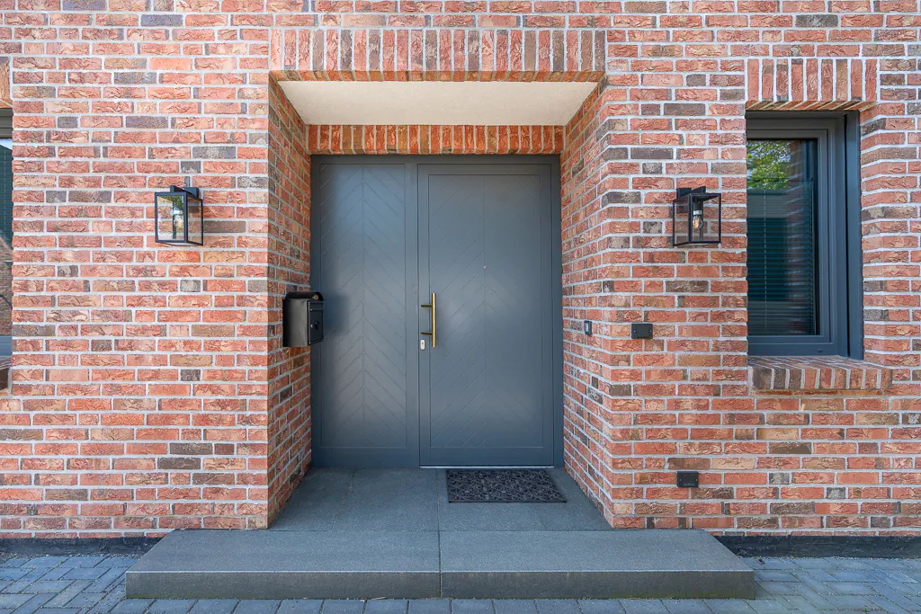 Red brick house facade with a gray front door, gold handle, and black mailbox. Two black lanterns flank the door.