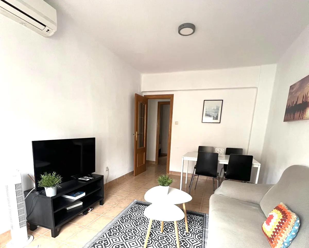Bright living room with a beige sofa, black TV stand, two white coffee tables, and a dining area with black chairs.