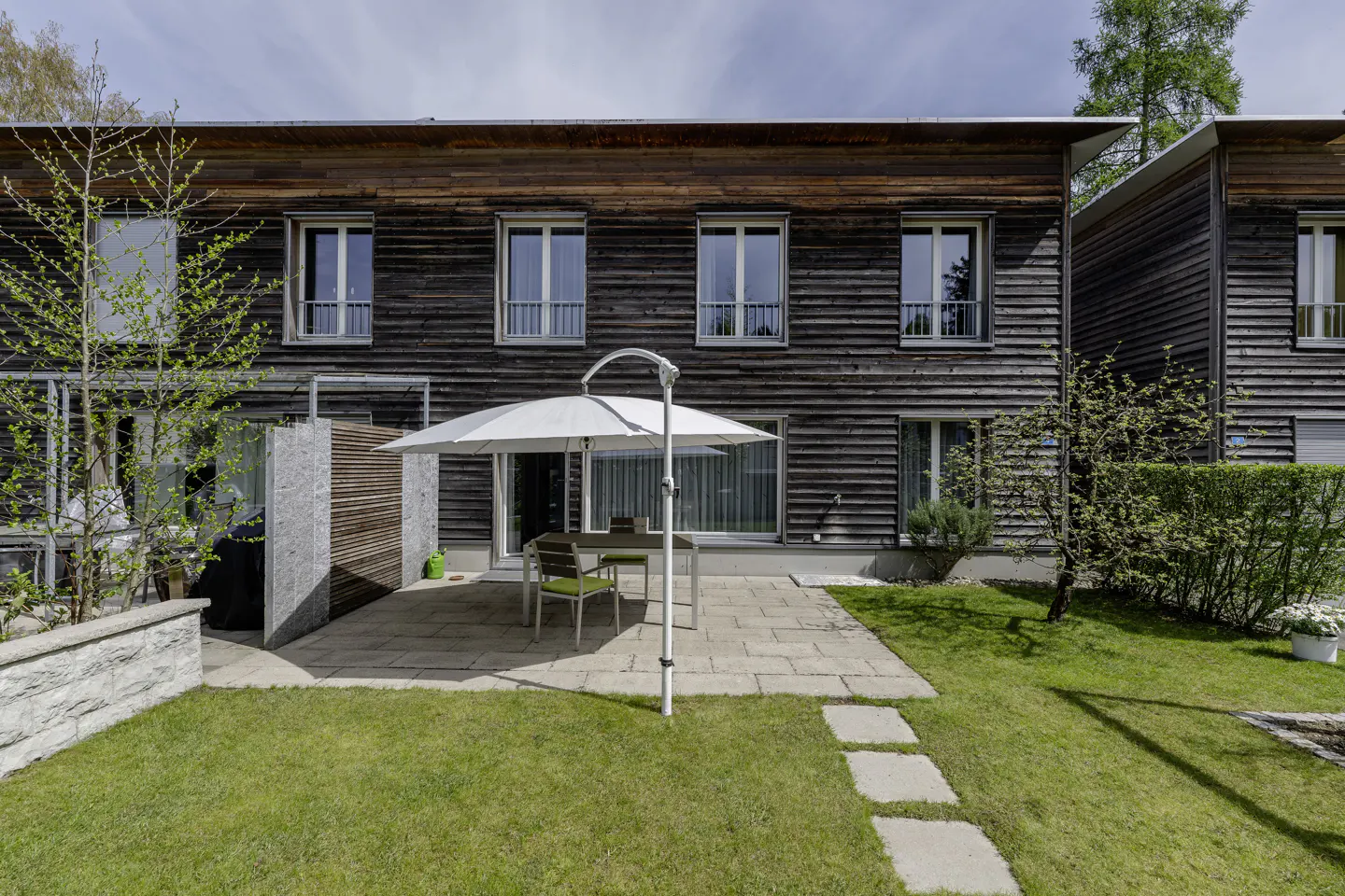Exterior of a wood-clad house with a patio, table, chairs, and a white umbrella. A green lawn and stone path are visible.