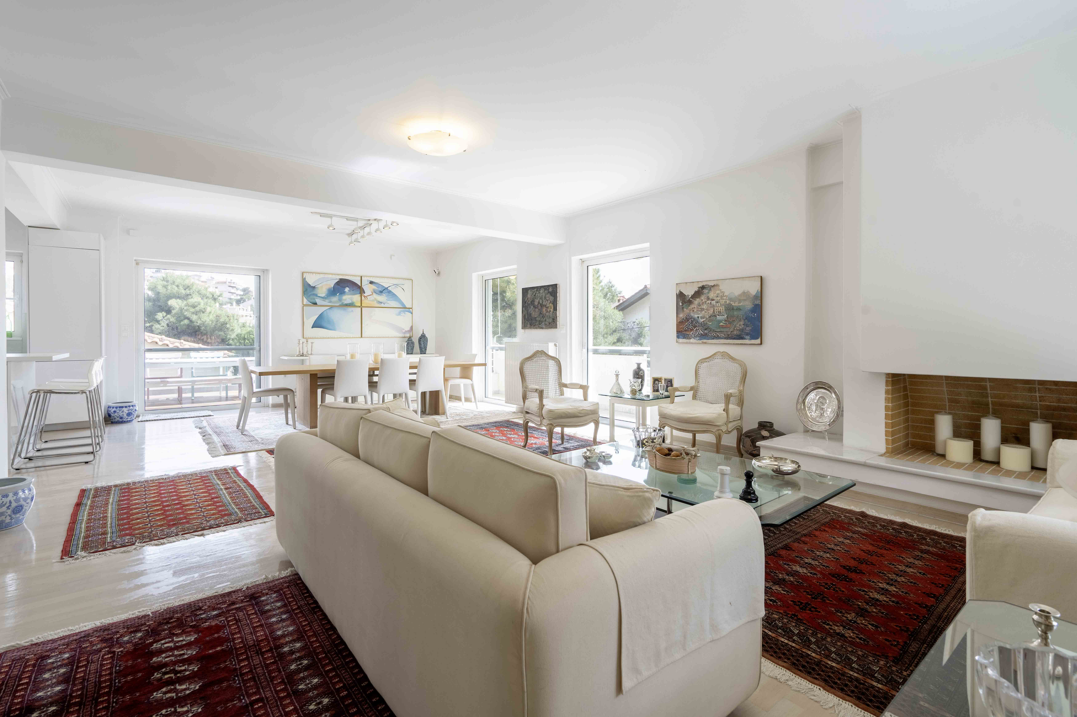 Bright, open living room with white walls, light wood floors, and red rugs. Cream sofas and chairs surround a glass coffee table. Dining area in background.