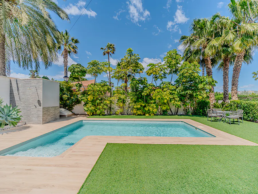 Outdoor pool with light blue water, surrounded by a light wood deck and green lawn, with palm trees and blue sky.