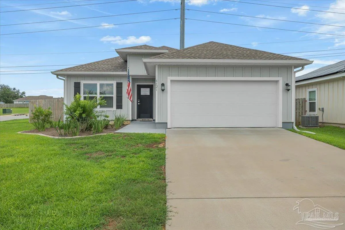 Single-story gray house with a two-car garage, front lawn, and American flag by the front door under a blue sky.