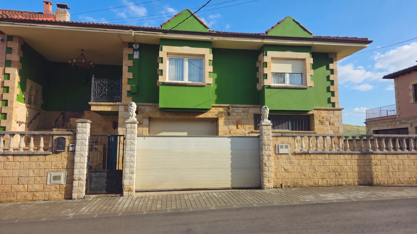 Two-story green house with stone accents, a white garage door, and a black iron gate. Lion statues adorn the gate posts.