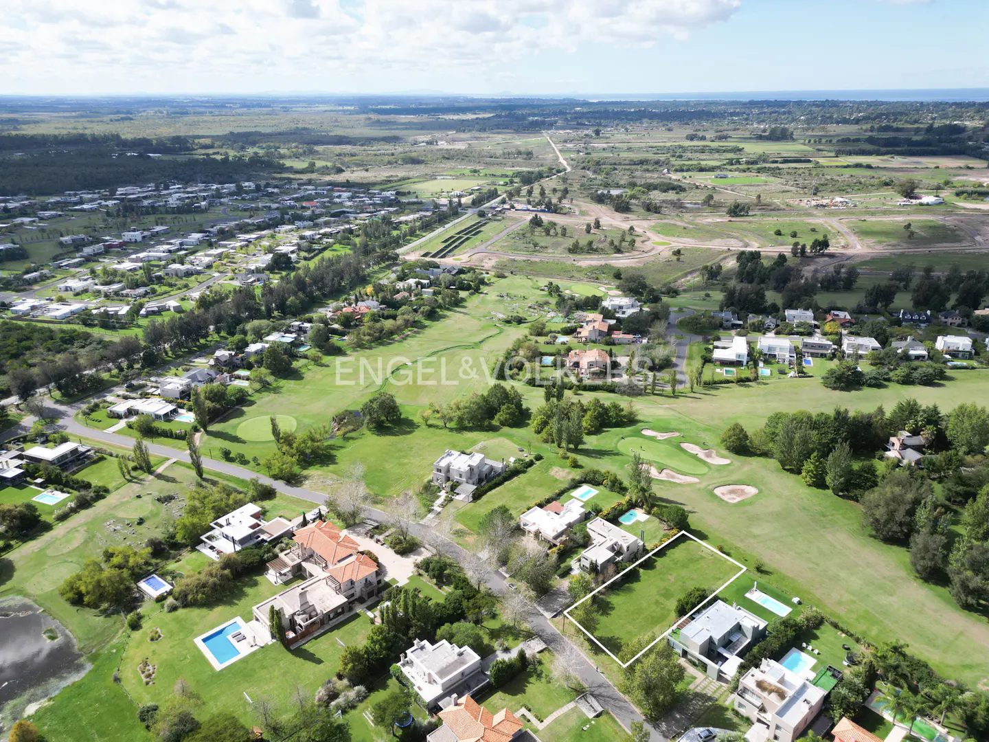 Aerial view of a green lot outlined in white, surrounded by houses, trees, and a golf course. Distant landscape under a blue sky with clouds.