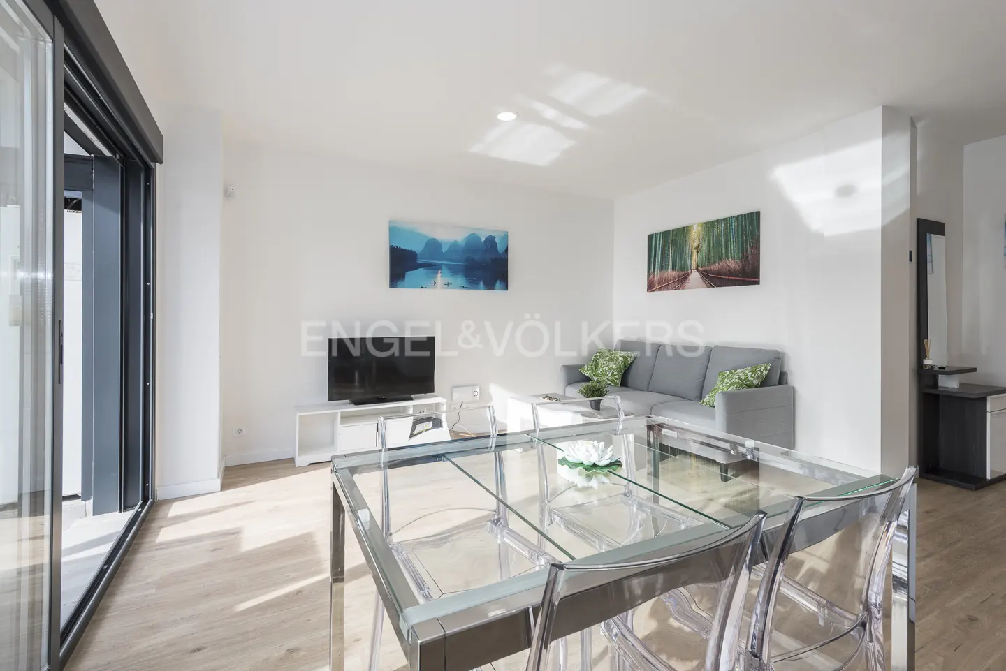 Bright living room with a glass table, clear chairs, gray sofa, and a TV. Artwork adorns the white walls.
