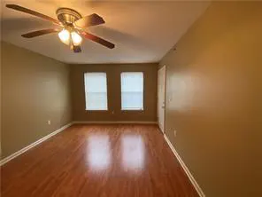 Empty room with wood floors, tan walls, and a ceiling fan with lights. Two windows with white blinds are visible.