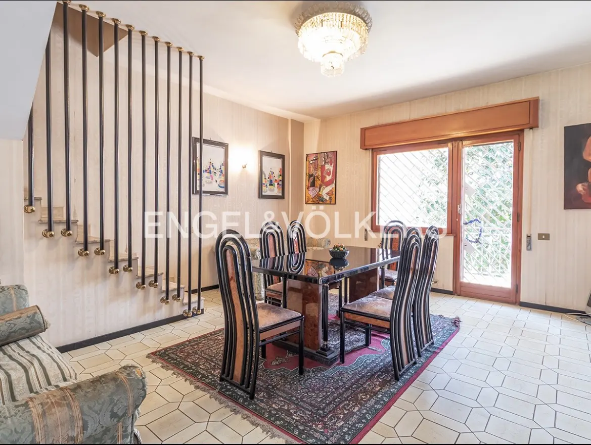 A dining room with a black table, six chairs, and a chandelier. A staircase with black railings is on the left.
