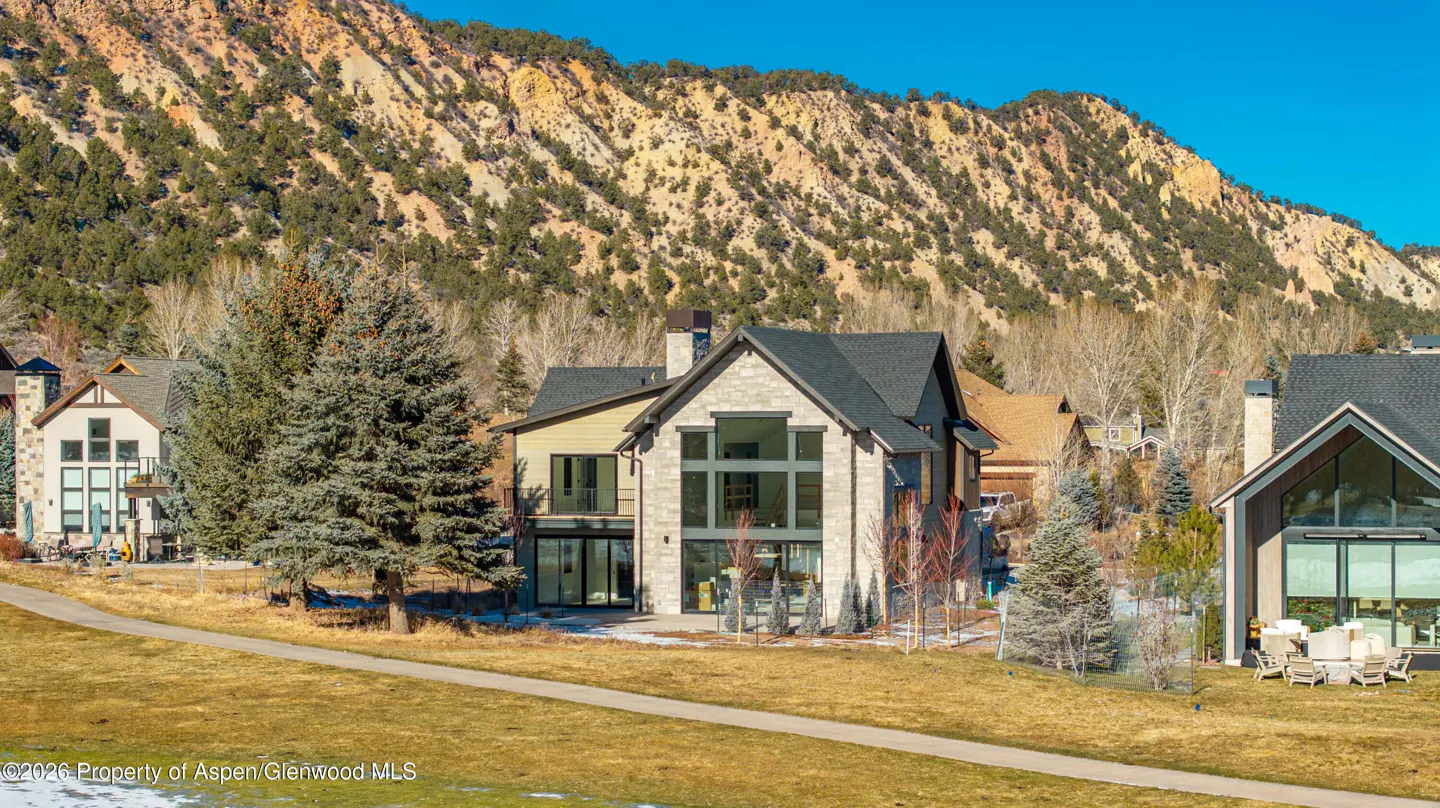 A modern two-story home with stone and siding, large windows, and a dark roof, set against a mountain backdrop.