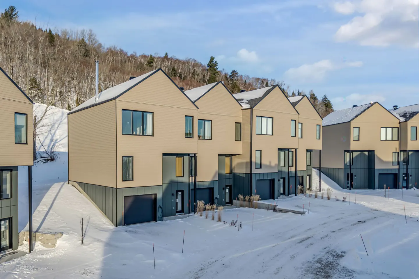Row of modern townhouses with beige siding and dark trim, set in a snowy landscape with a wooded hill in the background.