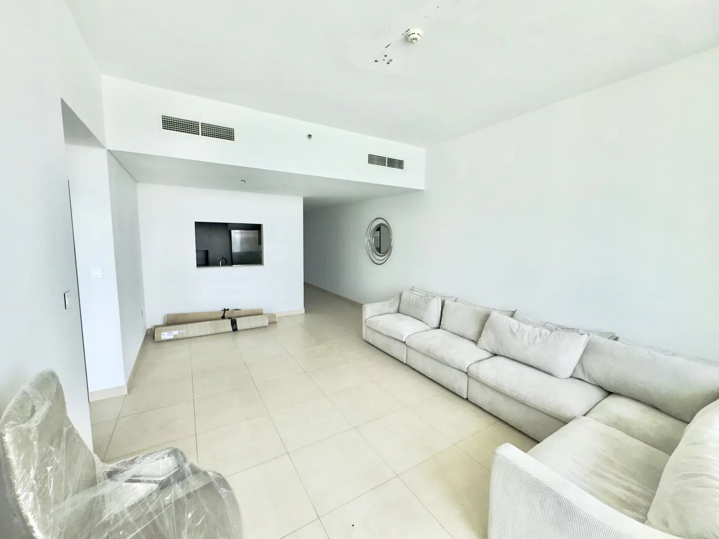 Bright, empty living room with white walls, tile floor, and a large, light gray sectional sofa. A chair is wrapped in plastic.