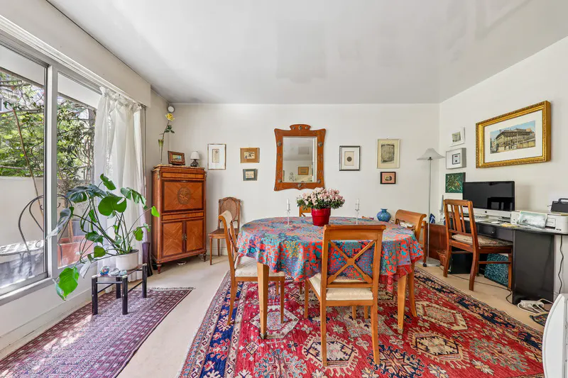 A bright dining room with a round table covered in a colorful tablecloth, surrounded by wooden chairs on a red patterned rug. A desk with a computer is in the background.
