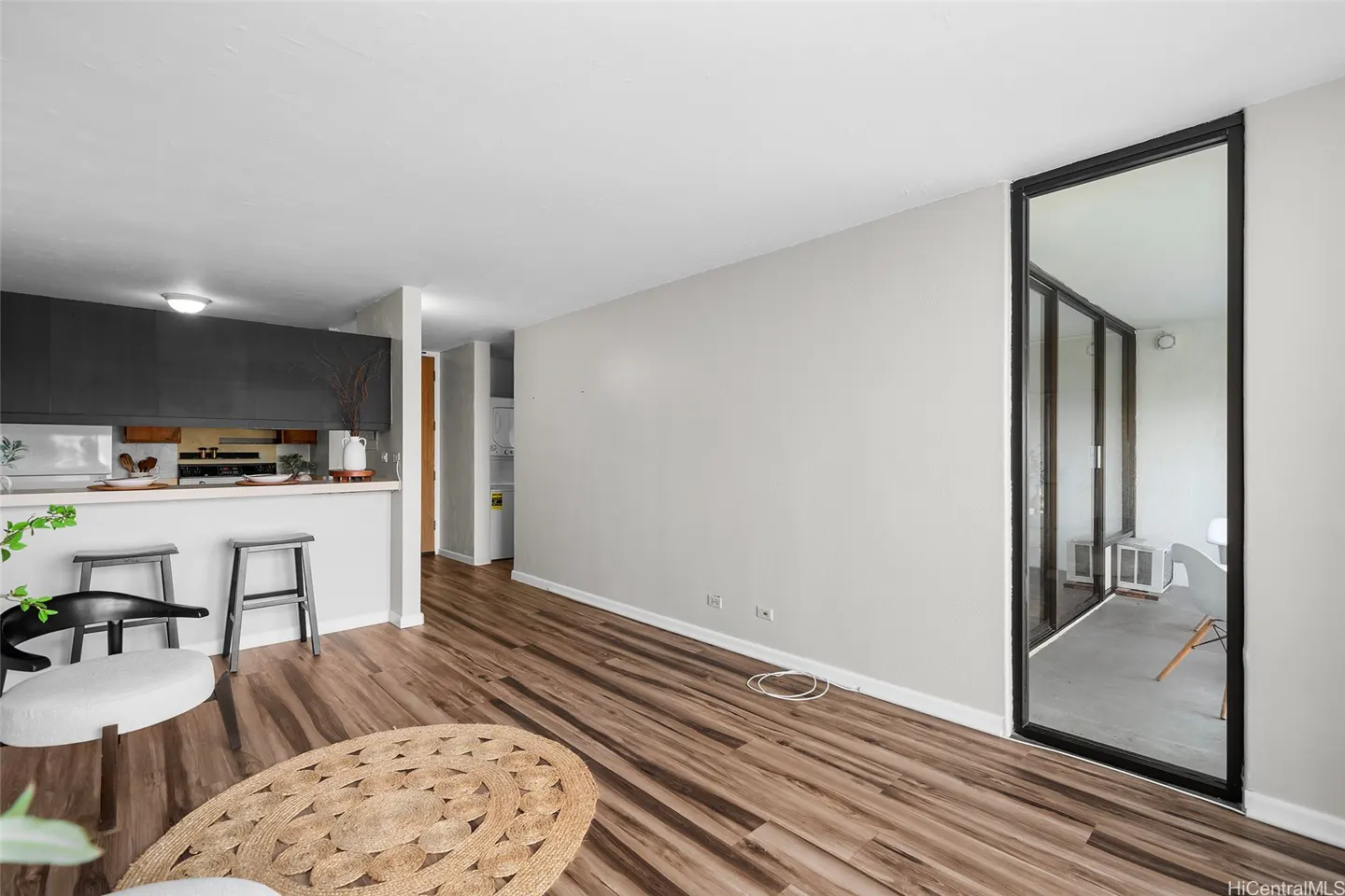 A bright living room with wood floors, a jute rug, and a kitchen bar with stools. A black-framed glass door leads to a balcony.