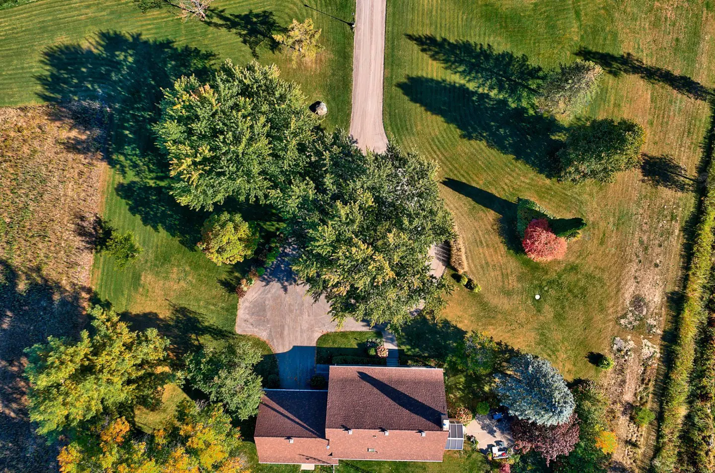Aerial view of a house with a brown roof, surrounded by green trees and grass. A driveway leads to the house.