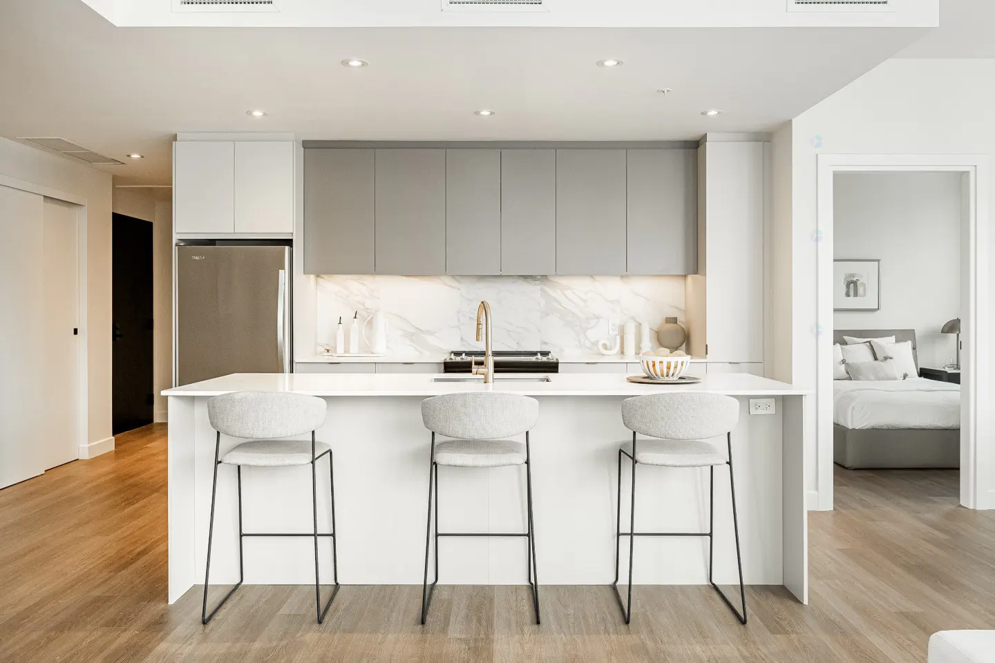 Bright, modern kitchen with white island, three gray stools, stainless steel fridge, and gray cabinets. Bedroom visible through doorway.