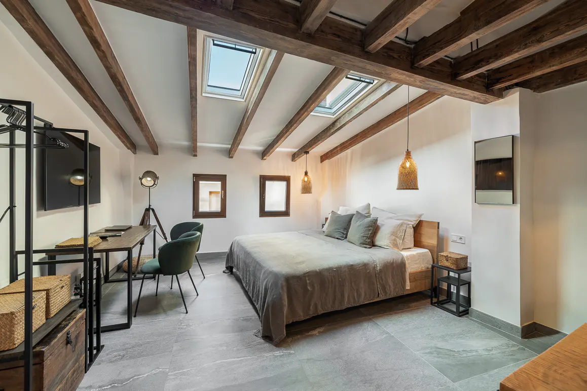 Bedroom with exposed beams, skylights, and gray tile floor. A bed with gray bedding and pillows is centered. A desk and chairs are on the left.