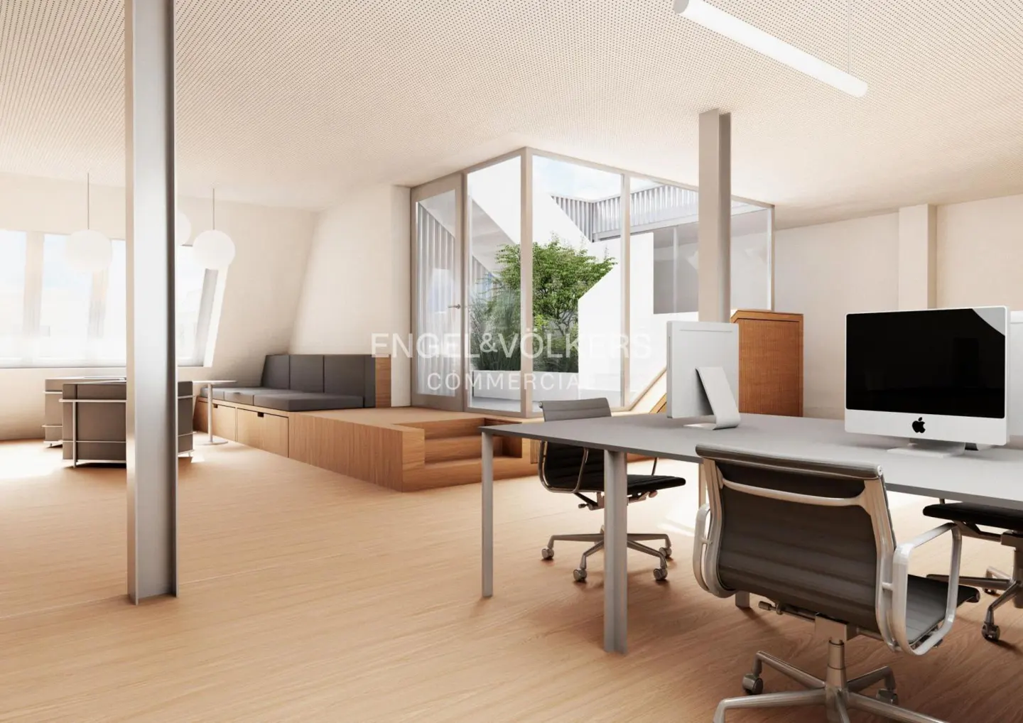Bright office space with desks, chairs, and computers. A gray sofa sits on a raised wooden platform near a glass-enclosed courtyard.