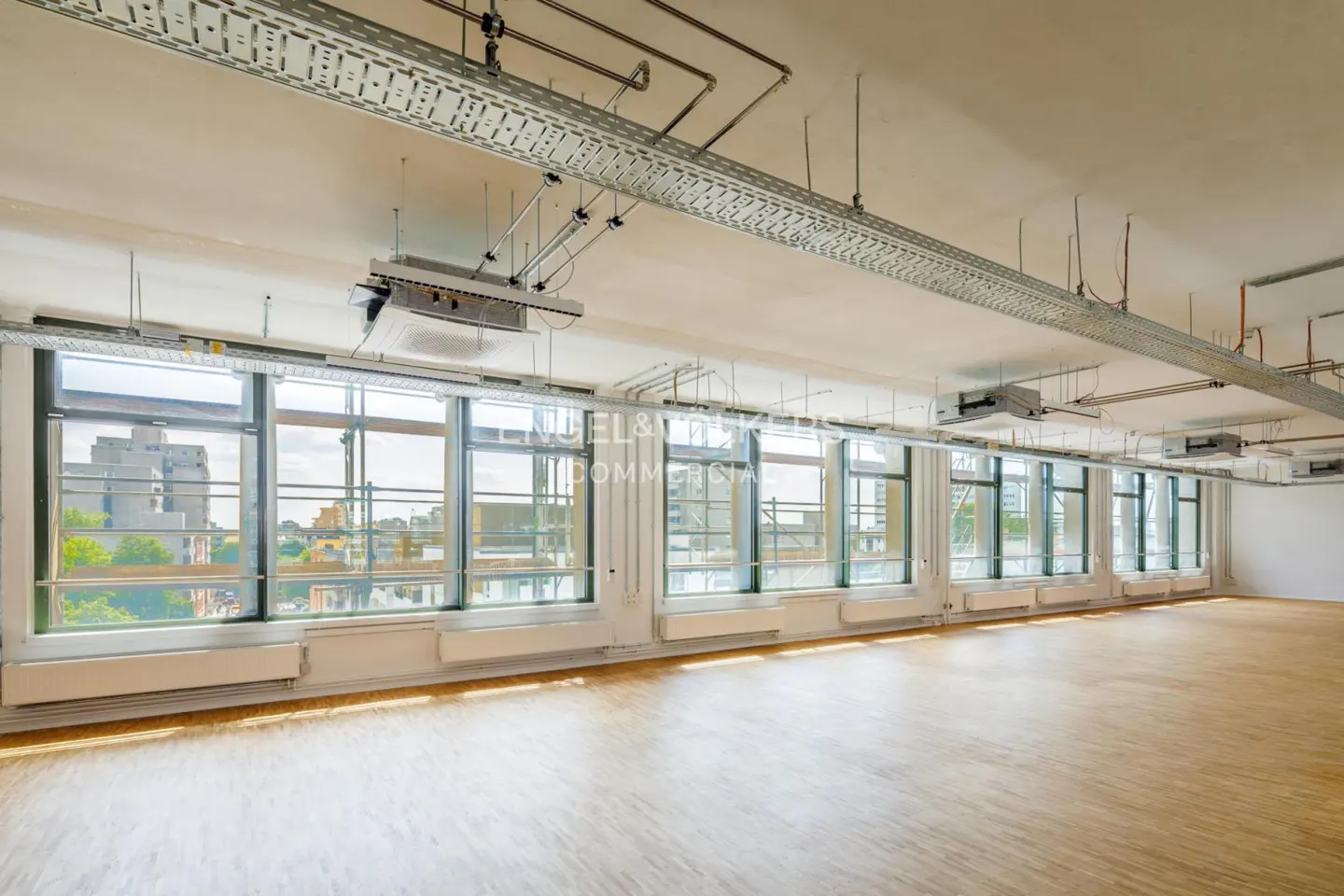 Bright, empty office space with wood floors and large windows overlooking a city. Metal cable trays run along the ceiling.