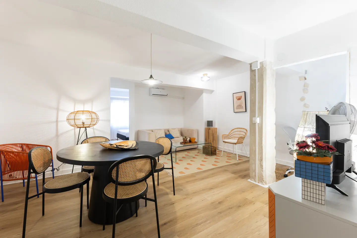 Bright, open-concept living space with a black dining table, rattan chairs, and a beige sofa. Wood floors and white walls create a modern, airy feel.