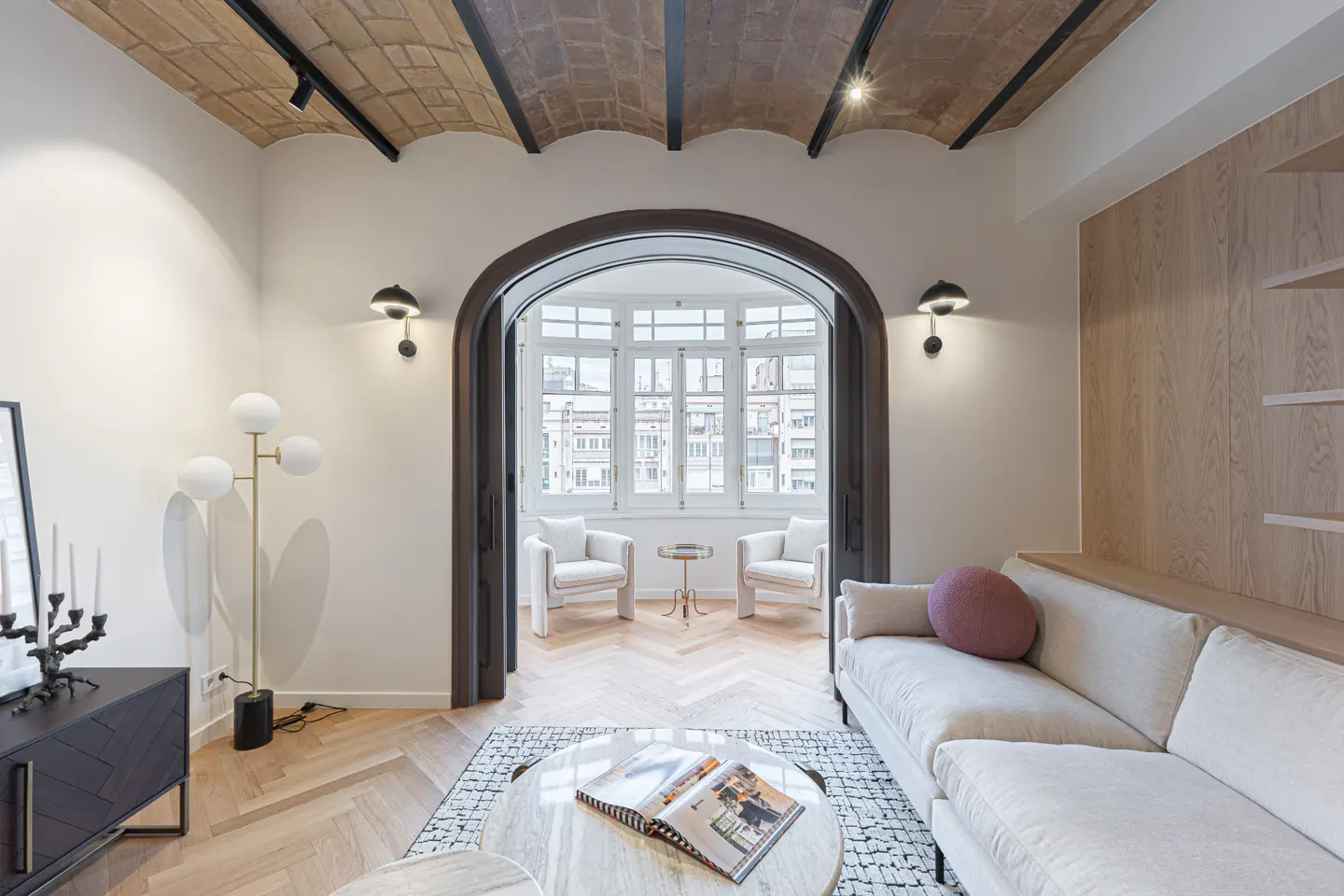 Bright living room with herringbone wood floors, arched doorway to a sunroom, and a white sofa with a pink pillow.