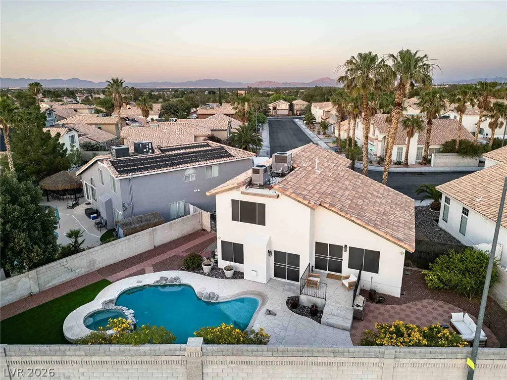 Aerial view of a white two-story house with a tile roof, backyard pool, spa, and patio in a desert neighborhood at sunset.