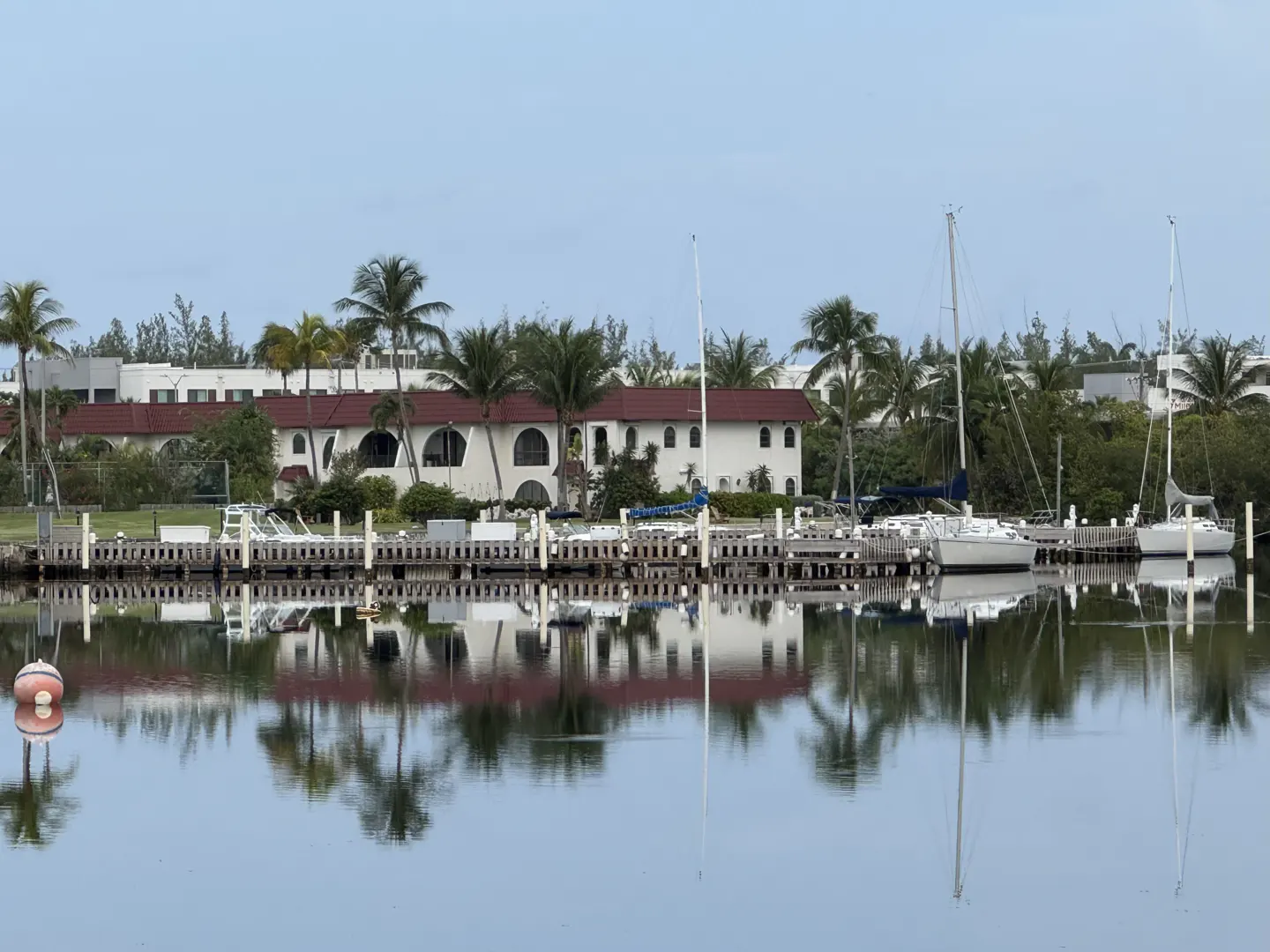 Waterfront view of a white building with a red roof, palm trees, and sailboats docked at a pier, all reflected in the water.