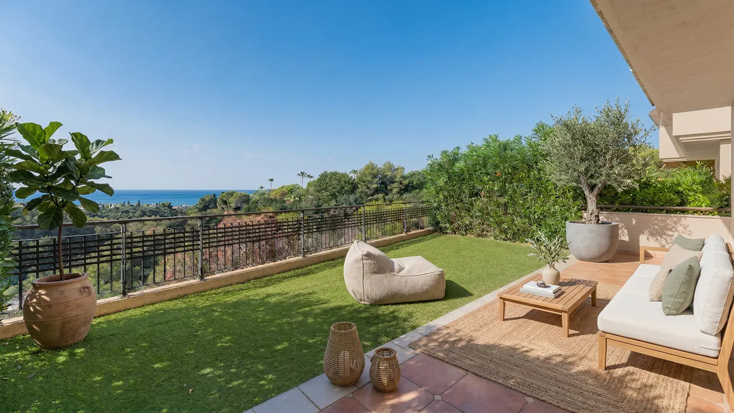 Outdoor patio with green lawn, furniture, and ocean view. A beige sofa, beanbag chair, and potted plants decorate the space.