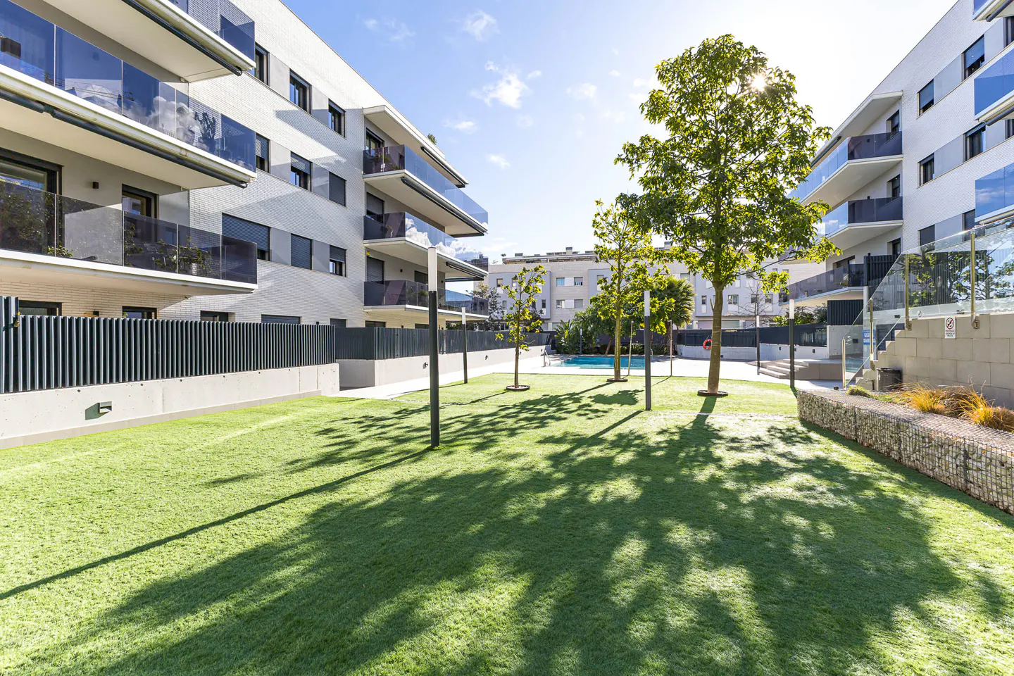 Apartment complex with a green lawn, trees, and a pool in the background on a sunny day.