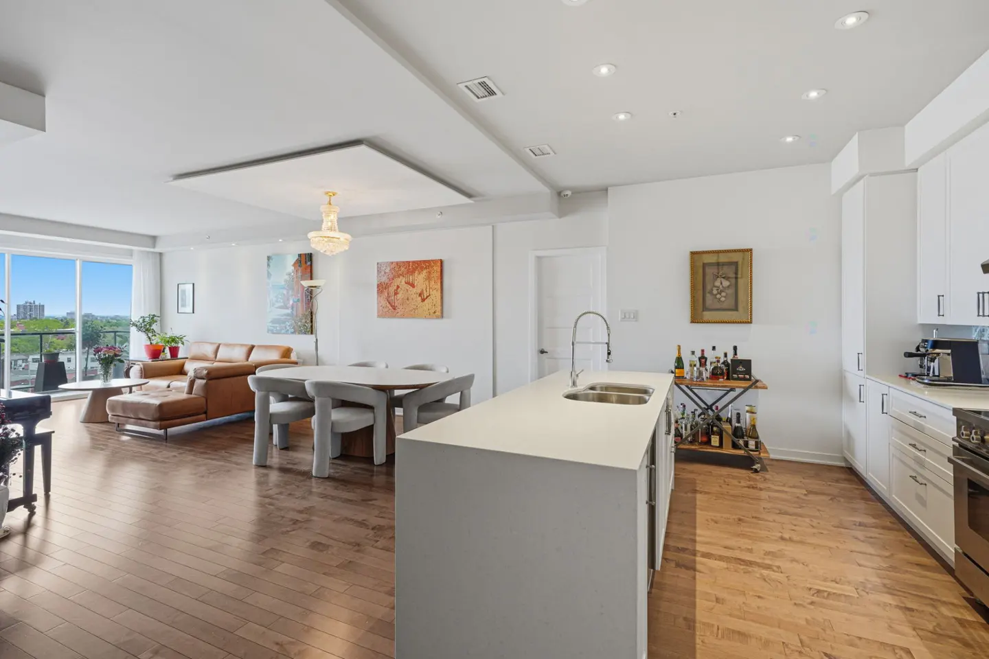 Bright, open-concept living space with hardwood floors. A brown leather sofa faces a kitchen with white cabinets and island. A dining table sits nearby.