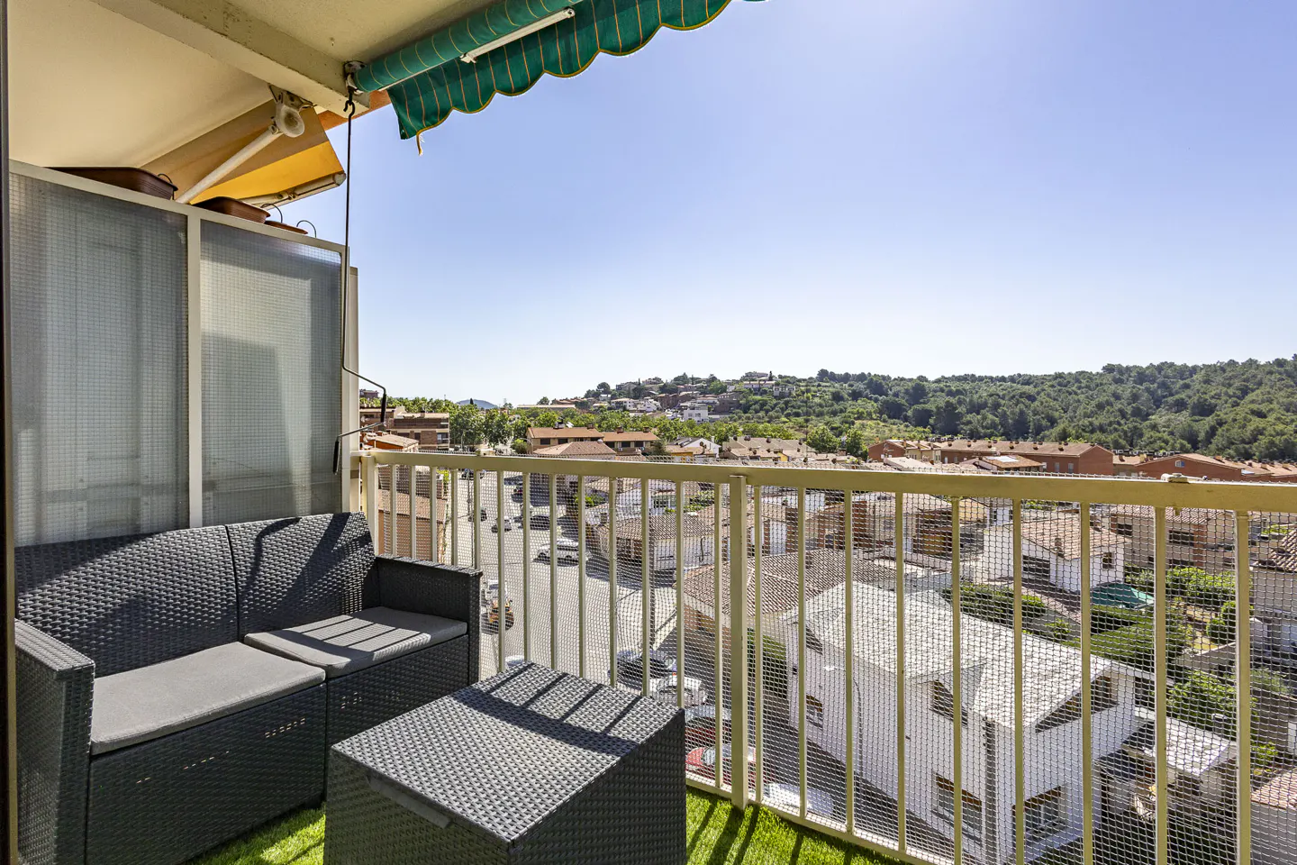 Balcony with black wicker furniture, green awning, and a view of a town with red-tiled roofs and green hills under a blue sky.