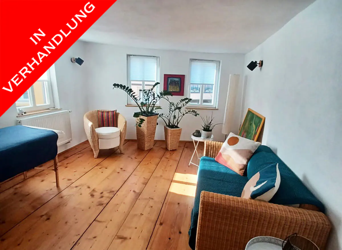 Bright living room with wood floors, white walls, and two windows. Wicker furniture, plants, and a blue couch add color. "In Verhandlung" banner.