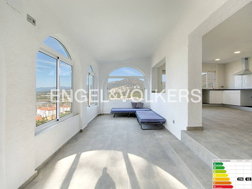 Bright, airy room with arched windows overlooking a town and mountains. Two blue daybeds sit near the windows, and a kitchen is visible in the background.