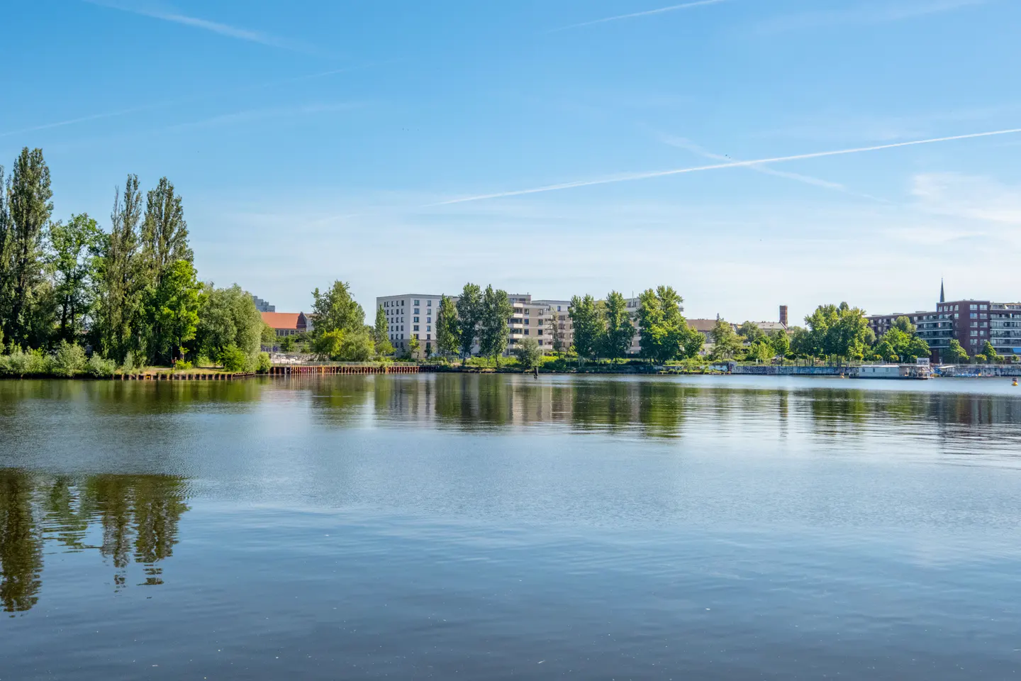 A calm river reflects the blue sky and surrounding green trees, with modern apartment buildings visible in the distance.