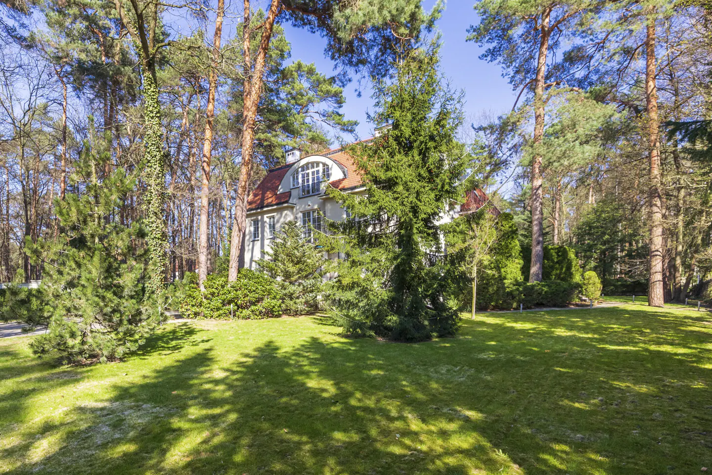 A white house with a red roof is surrounded by tall trees and a green lawn.