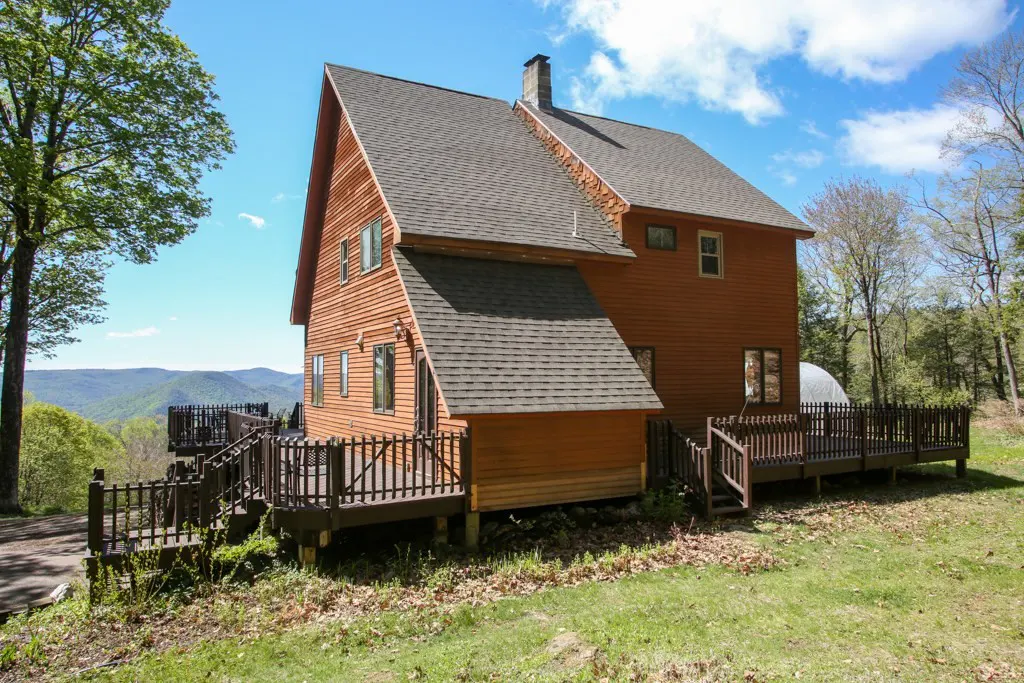 Exterior view of a two-story wood house with a deck, brown roof, chimney, and mountain views under a blue sky.