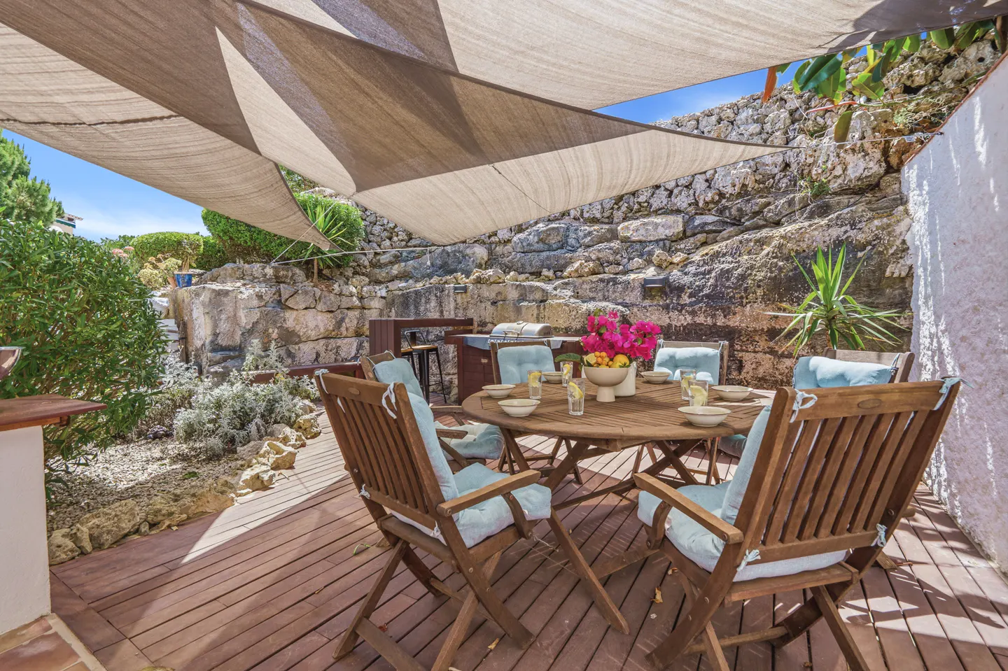 Outdoor patio with wooden furniture, light blue cushions, and a round table under a large beige sun shade against stone walls.