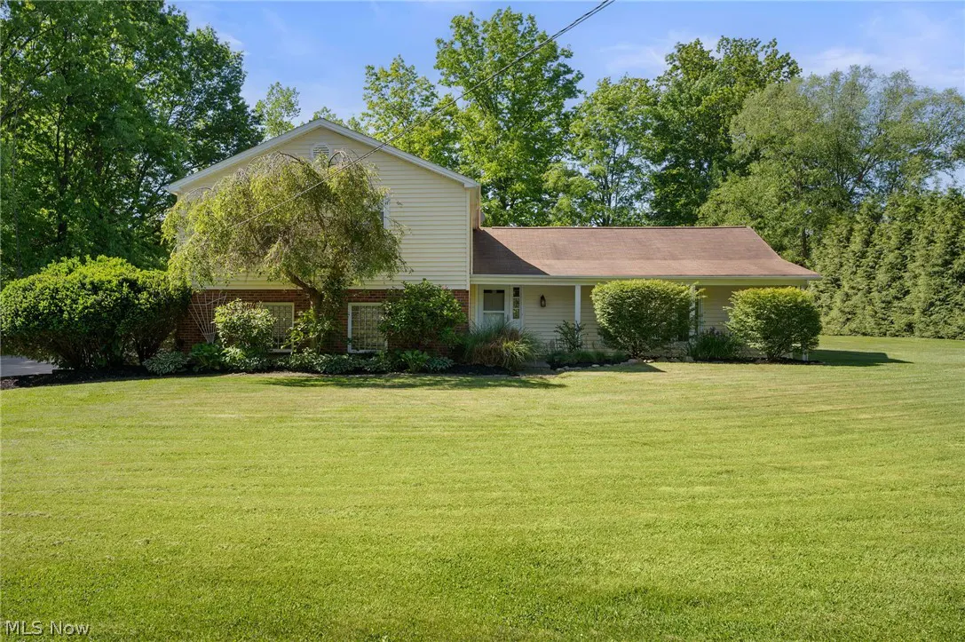 Two-story beige house with a brown roof, surrounded by lush green trees and a large, well-maintained front lawn.