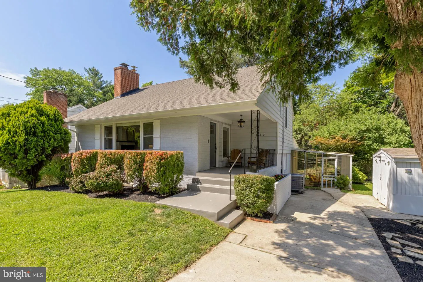 Exterior view of a light gray house with a brick chimney, a gray roof, and a small front porch. Green bushes line the front.