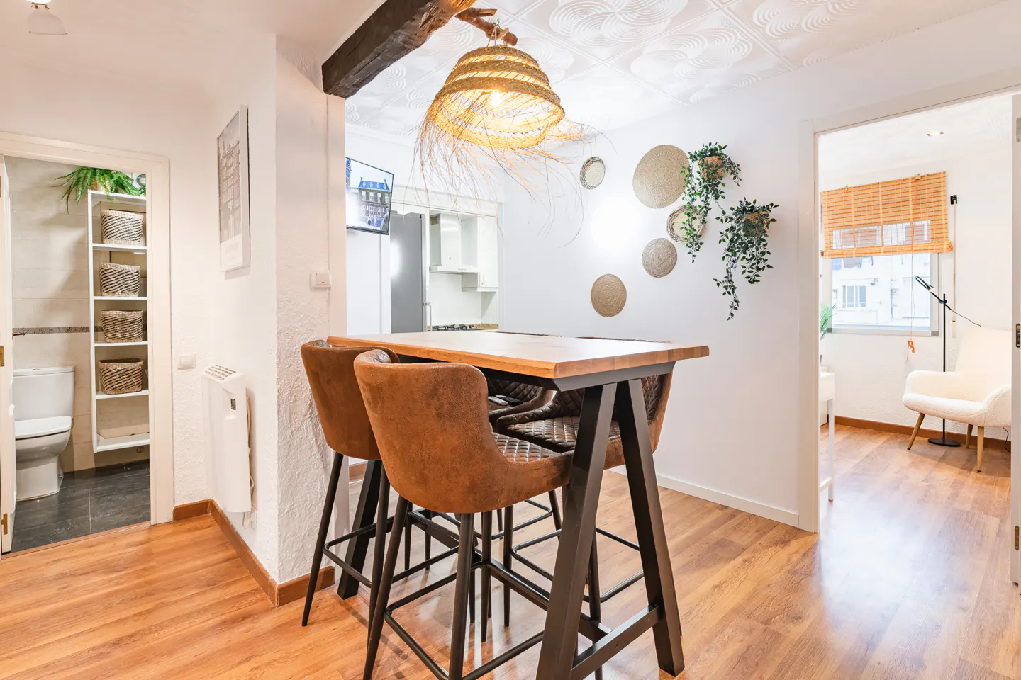 Interior view of a modern apartment with a wooden floor, a high table with brown chairs, and a straw lamp. There is a bathroom and a living room in the background.