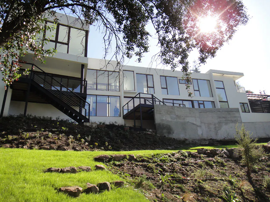 Modern white house with large windows and black metal stairs on a grassy hill under a sunny sky.