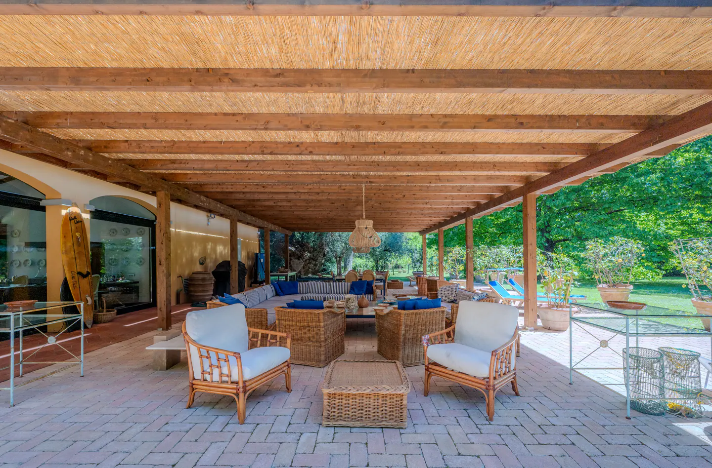 Outdoor patio with wicker furniture under a wood pergola with a thatched roof. Lush green trees in the background.