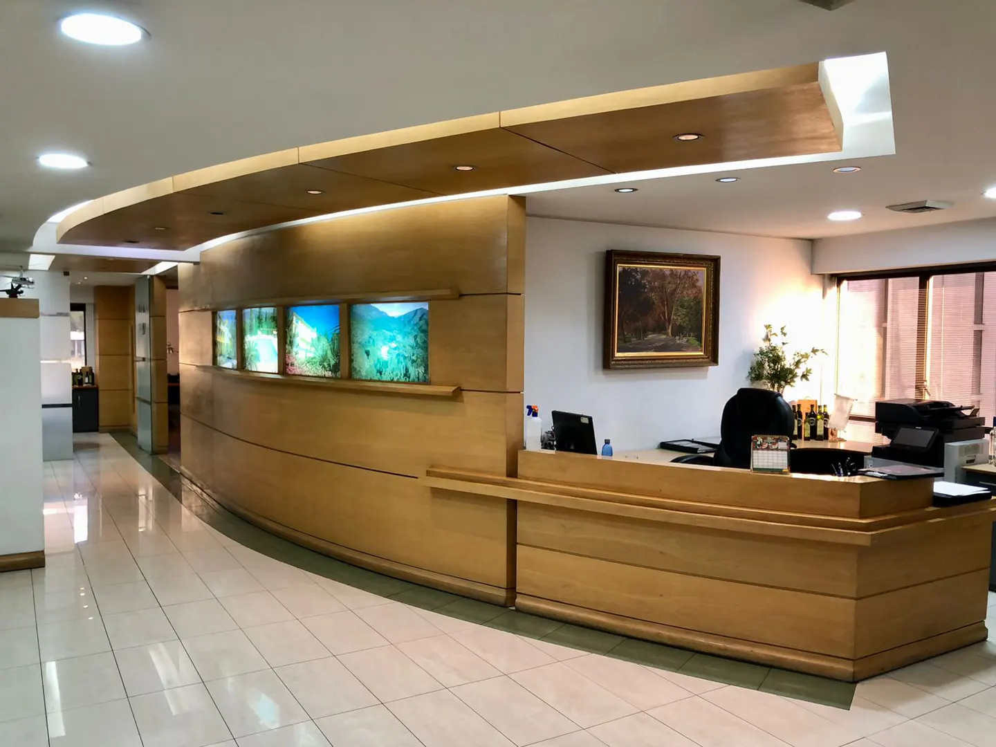 Office reception area with a curved wooden desk, landscape art, and white tile floor.