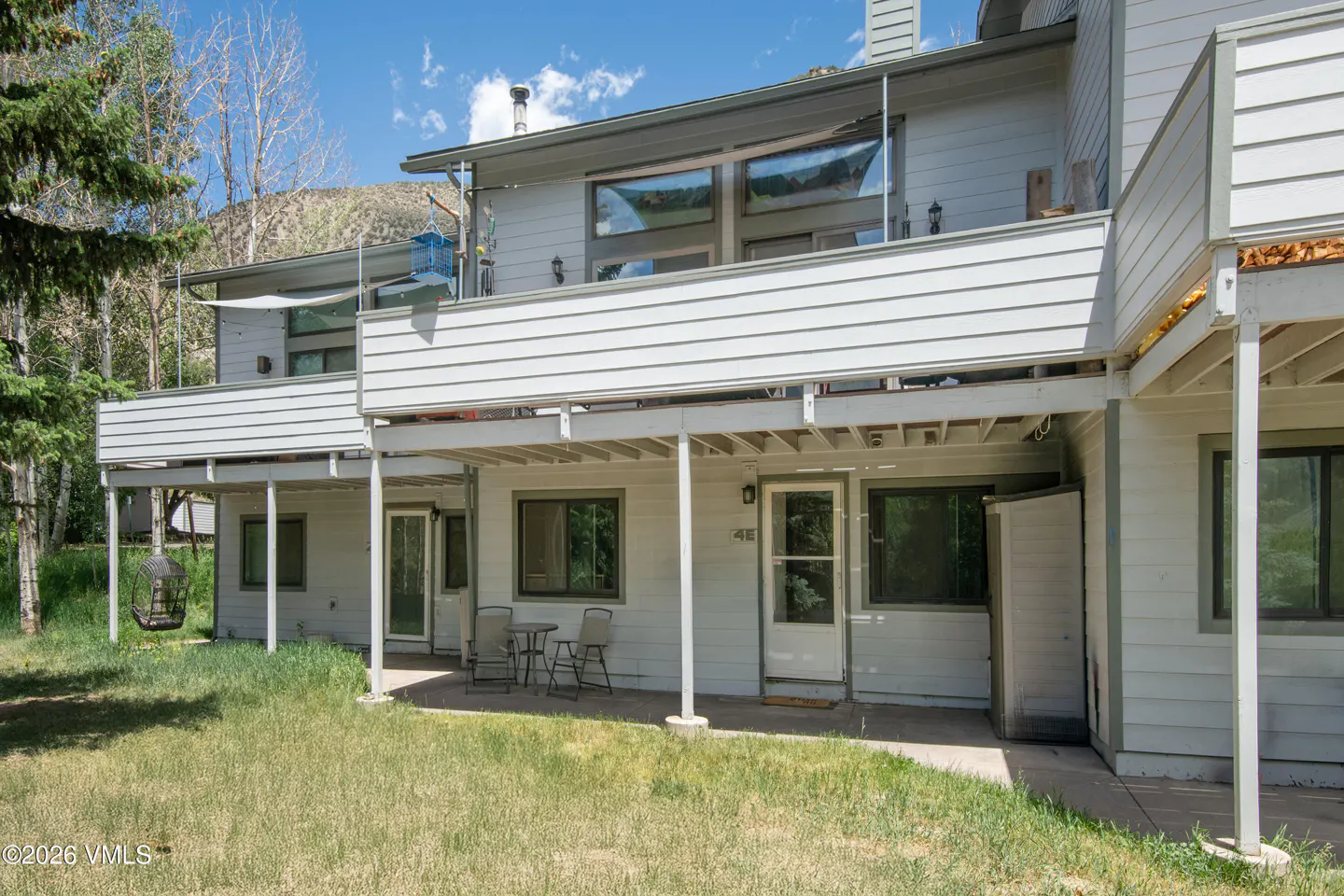 Back exterior of a two-story light gray home with a deck, patio, outdoor seating, and a grassy yard against a mountain.