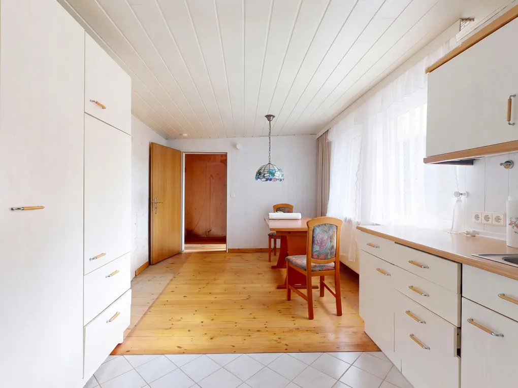 Bright kitchen with white cabinets, wood floors, and a dining table with floral chairs. An open doorway leads to another room.